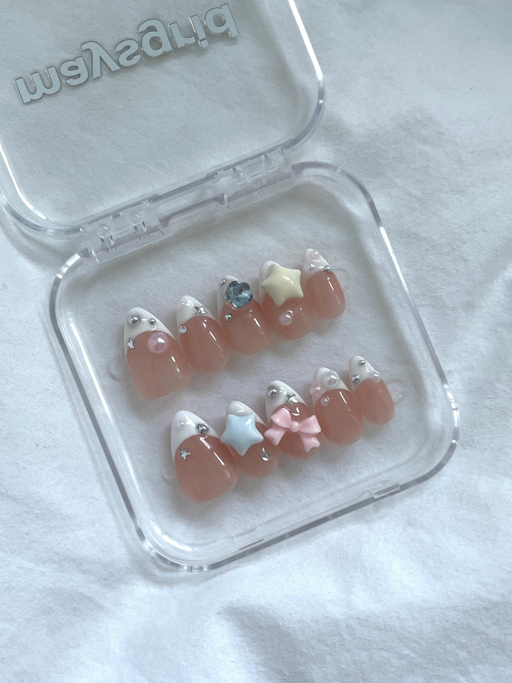 A set of false press-on nails displayed in a clear plastic case on a white fabric background. The nails feature a glossy peachy-pink base with white tips, some decorated with small rhinestones, and one pink bow accent.