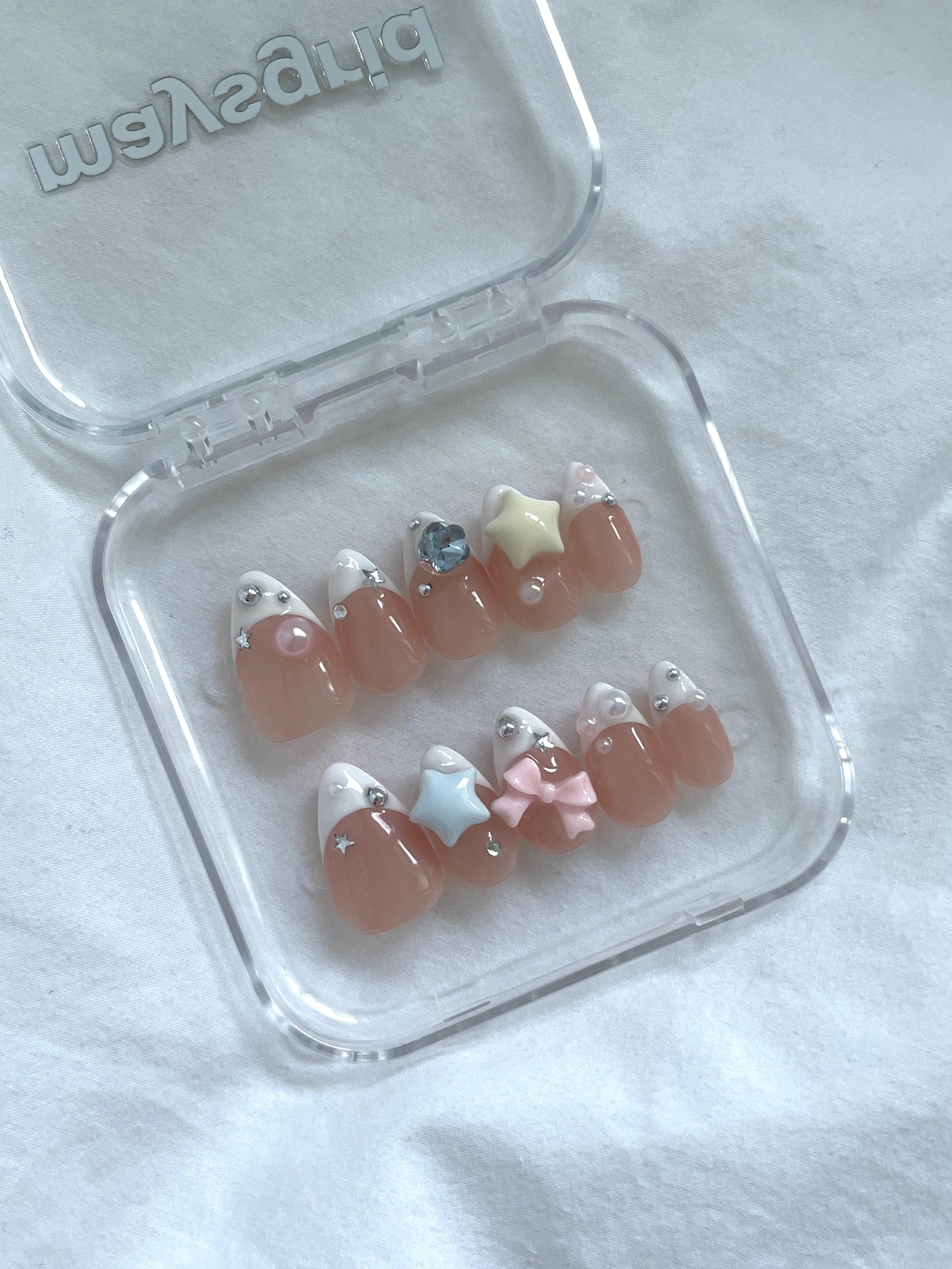 A set of false press-on nails displayed in a clear plastic case on a white fabric background. The nails feature a glossy peachy-pink base with white tips, some decorated with small rhinestones, and one pink bow accent.