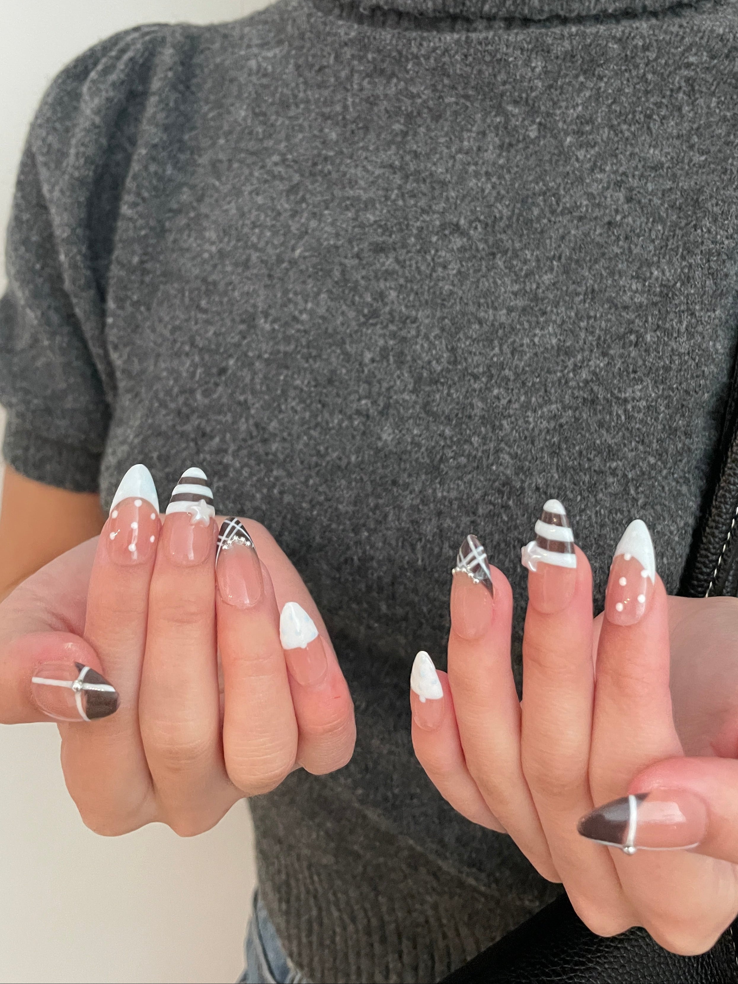 Close-up of a set of espresso brown and baby blue french tip press on nails in a clear plastic case on a white background. White and blue polka dot detailing, stripes on middle finger, and plaid designs on ring fingers. Pearl star and silver gem detailing—gray turtleneck background. 