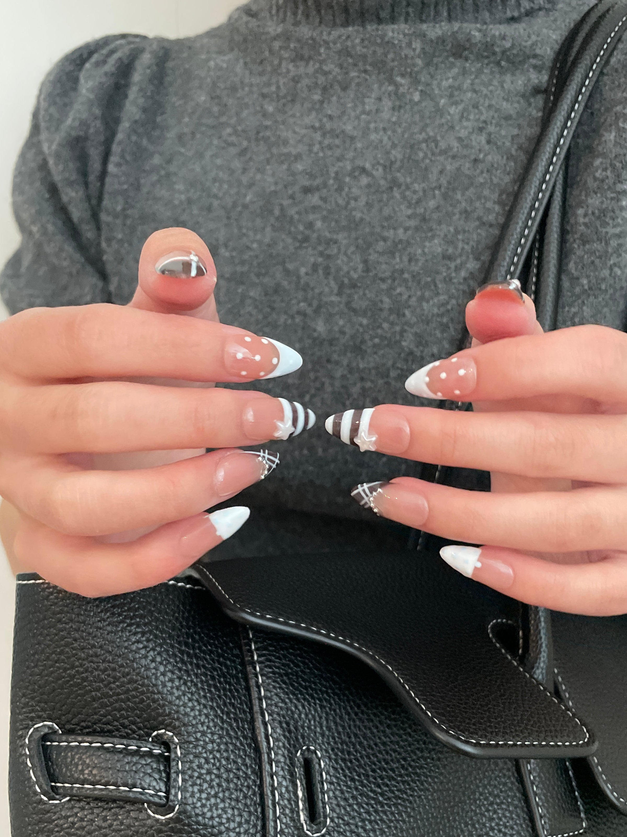 Close-up of a set of espresso brown and baby blue french tip press on nails in a clear plastic case on a white background. White and blue polka dot detailing, stripes on middle finger, and plaid designs on ring fingers. Pearl star and silver gem detailing—gray turtleneck background. Black shoulder bag on bottom of picture