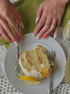 Slice of cake on a plate with hands holding a fork, green placemat underneath. Set of press on nails in orange, pink, pale yellow, and pale pink. Silver and pearl detailing. 