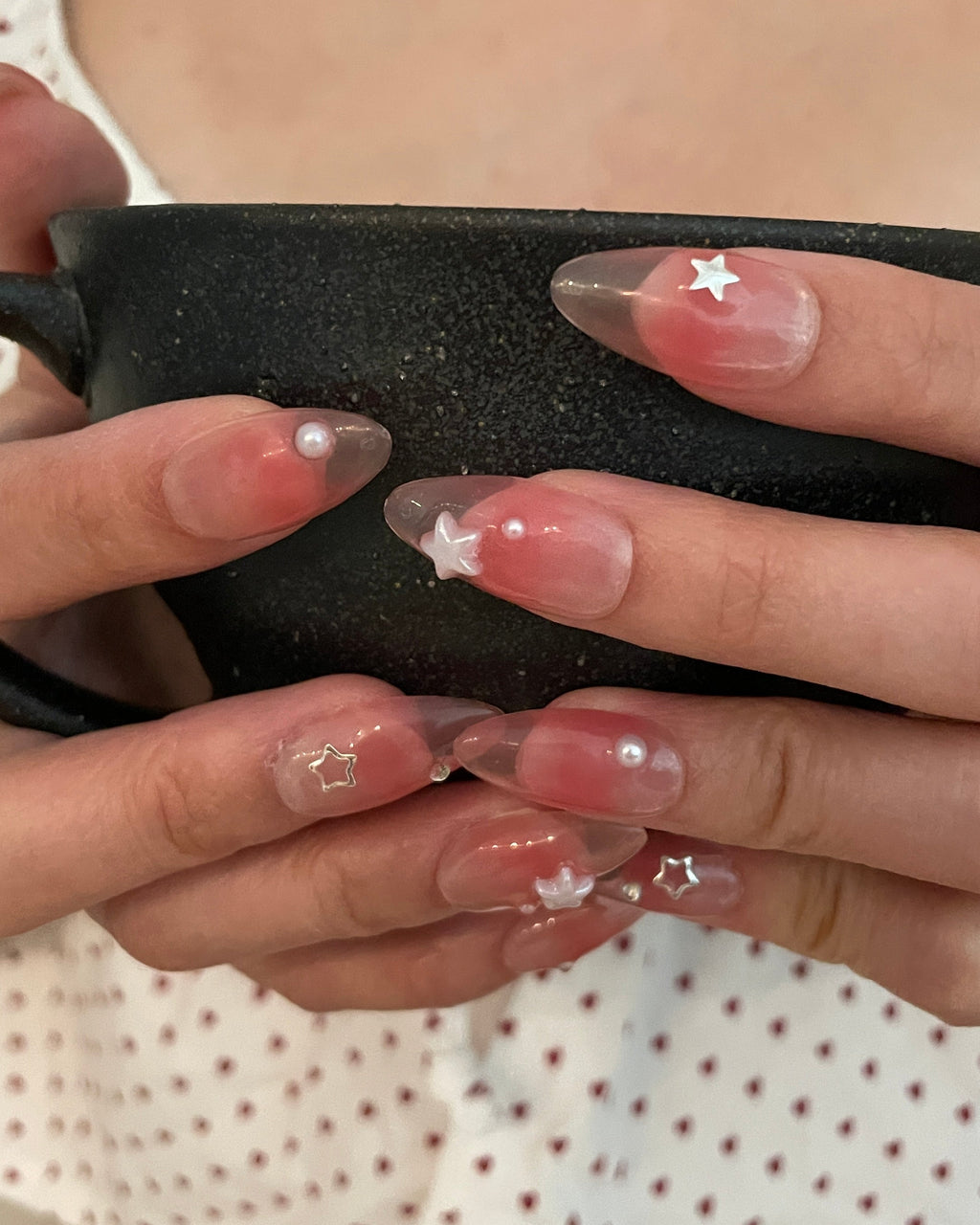 Close-up of hands with a set of jelly pink (translucent pink) press ons. Silver stars, silver gems, and pearl star, and pearl detailing, hands holding a black tea cup.