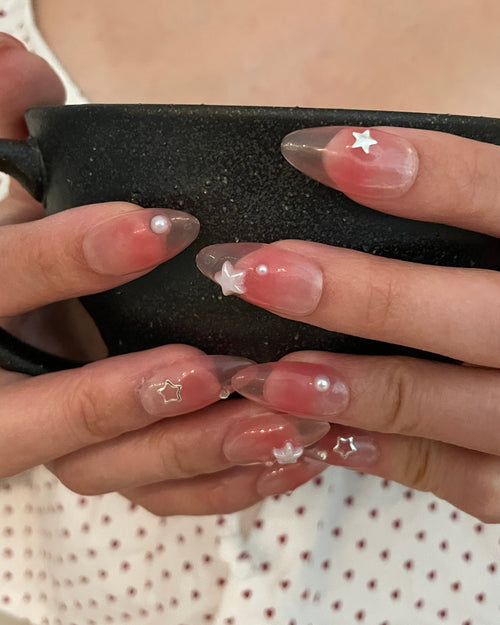 Close-up of hands with a set of jelly pink (translucent pink) press ons. Silver stars, silver gems, and pearl star, and pearl detailing, hands holding a black tea cup.